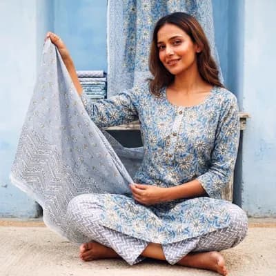 A woman in a blue printed kurta set sitting in daylight against a blue wall, holding out her dupatta